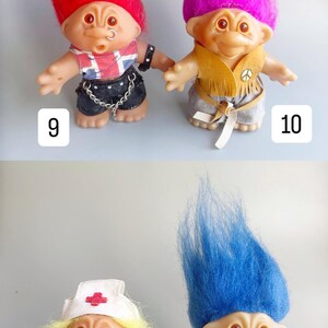 May include: Four troll dolls with different colored hair and outfits. The first doll has red hair and is wearing a black leather jacket with a Union Jack design, black pants, and a silver chain. The second doll has purple hair and is wearing a brown suede vest with fringe, gray pants, and a peace sign. The third doll has blonde hair and is wearing a blue nurse's uniform with a red cross. The fourth doll has blue hair and is wearing a white dress with lace.