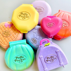 May include: A collection of vintage Polly Pocket toys in various colors and shapes, including a heart-shaped compact, a suitcase, a clam shell, a horse-shaped compact, and a quilted compact. Each compact features the Polly Pocket logo with a heart.
