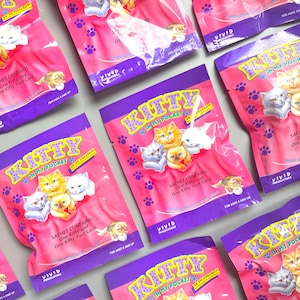 May include: Pink Kitty in My Pocket collectible sachets with purple borders. Each sachet features illustrations of kittens in various colors. The text on the sachets reads "Kitty in My Pocket" and "25 to Collect!" with the words "Sachet Contains: One Kitty and One Kitty Fact Card."