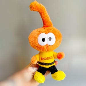 Puede incluir: Juguete de peluche naranja con ojos blancos grandes y una antena curva. El juguete lleva una camiseta amarilla con la palabra "SNORKS" en letras rojas, pantalones negros y pies amarillos. El juguete se sostiene en una mano.
