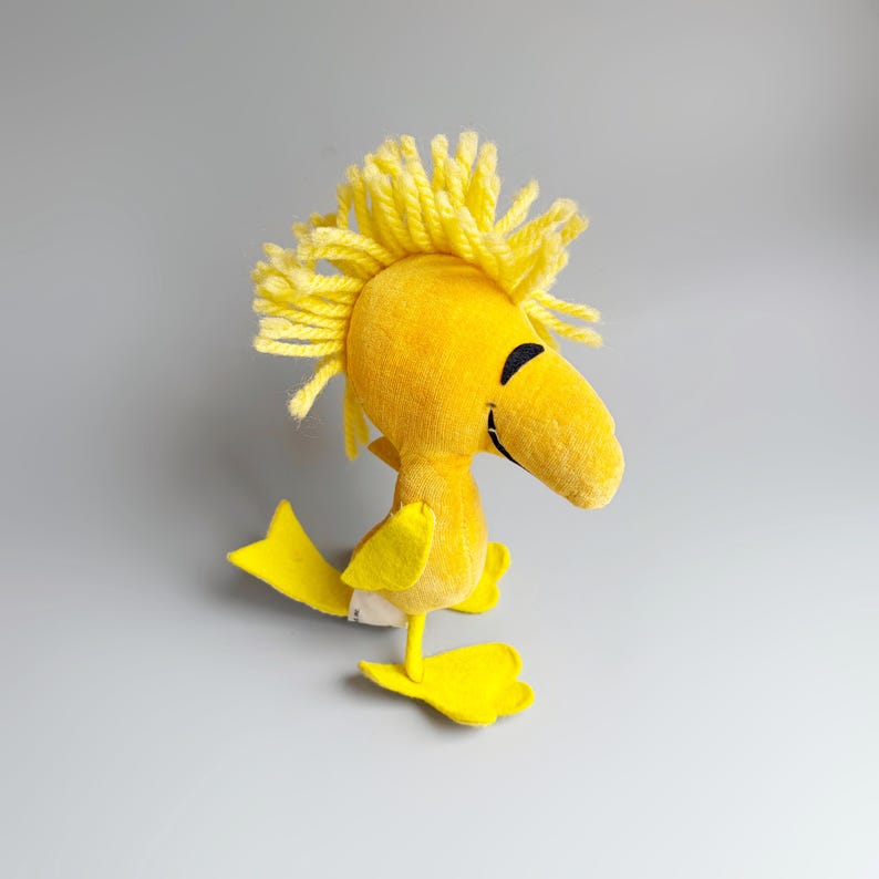Puede incluir: Un personaje de p&aacute;jaro de peluche amarillo con un pico grande y ojos negros. El p&aacute;jaro tiene un mech&oacute;n de hilo amarillo en la cabeza y est&aacute; de pie sobre dos pies amarillos. El personaje es de la tira c&oacute;mica Peanuts.