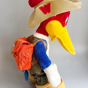 May include: A plush Woody Woodpecker toy wearing a red hat, tan cowboy hat with a red "W", and an orange backpack. The bird has a yellow beak, blue arms, and a green vest. The toy is wearing tan shoes.