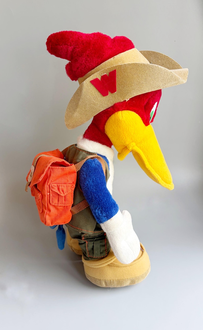 May include: A plush Woody Woodpecker toy wearing a red hat, tan cowboy hat with a red "W", and an orange backpack. The bird has a yellow beak, blue arms, and a green vest. The toy is wearing tan shoes.