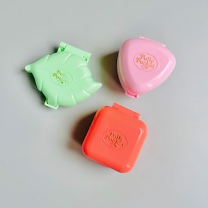 May include: Three vintage Polly Pocket compacts in green, pink, and orange. The green compact is leaf-shaped, the pink is triangular, and the orange is rectangular. Each compact has the words "Polly Pocket" printed on it.