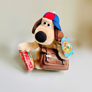 Può includere: Un cane di peluche marrone e bianco che indossa un berretto da baseball blu e rosso e una borsa a tracolla marrone. Il cane tiene un libro rosso con il testo "ELECTRONICS FOR DOGS". Il cane ha un'etichetta attaccata alla sua borsa a tracolla che dice "Wallace & Gromit Sound Dog Game".