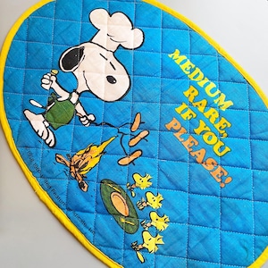 Może przedstawiać: Niebieska pikowana owalna podkładka pod garnek z Snoopy w czapce kucharskiej i napisem "MEDIUM RARE, IF YOU PLEASE!". Podkładka ma żółtą obwódkę i zawiera obrazy jedzenia i Woodstock.