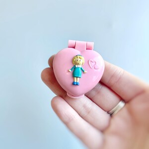 Puede incluir: Un medall&oacute;n de pl&aacute;stico rosa en forma de coraz&oacute;n con una peque&ntilde;a figura de mu&ntilde;eca de pl&aacute;stico en su interior. La mu&ntilde;eca lleva un vestido azul y tiene el pelo rubio.