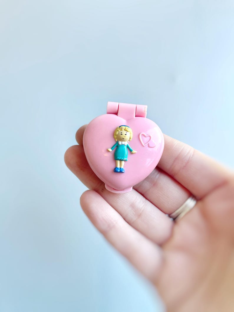 Puede incluir: Un medall&oacute;n de pl&aacute;stico rosa en forma de coraz&oacute;n con una peque&ntilde;a figura de mu&ntilde;eca de pl&aacute;stico en su interior. La mu&ntilde;eca lleva un vestido azul y tiene el pelo rubio.