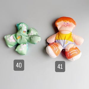Pu&ograve; includere: Due giocattoli di peluche vintage. Uno &egrave; un giocattolo verde a forma di croce con pois rosa. L'altro &egrave; una bambola con capelli arancioni, una camicia gialla e scarpe arancioni e bianche. I numeri 40 e 41 sono nell'immagine.