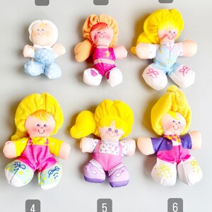 Pu&ograve; includere: Sei bambole di peluche vintage con volti dipinti e abiti colorati. Le bambole hanno capelli gialli o arancioni e indossano abiti nei toni del rosa, blu e viola. Ogni bambola ha un'espressione e un design unici.