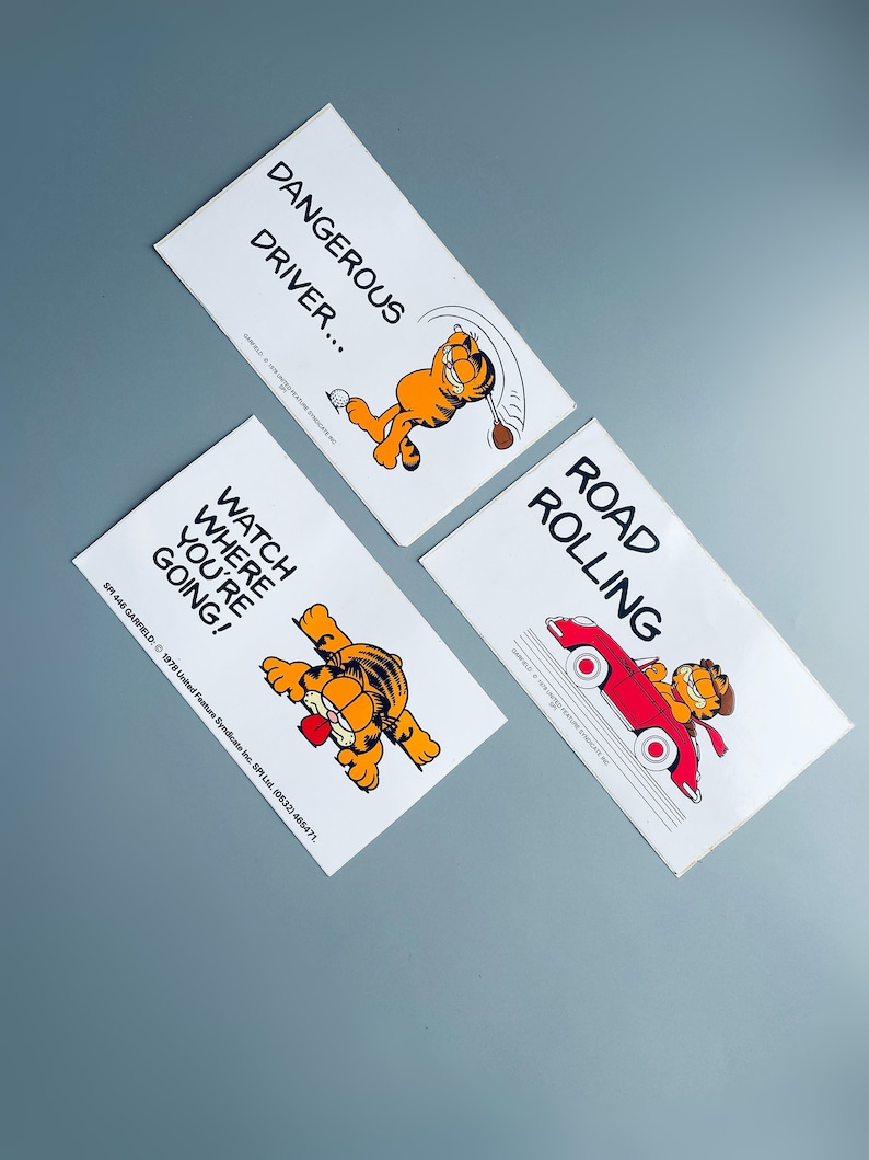 Puede incluir: Tres pegatinas de dibujos animados de Garfield blancas con diferentes lemas. La primera pegatina dice "Dangerous Driver..." y muestra a Garfield balanceando un palo de golf. La segunda pegatina dice "Watch Where You're Going!" y muestra a Garfield asom&aacute;ndose por detr&aacute;s de un cartel. La tercera pegatina dice "Road Rolling" y muestra a Garfield conduciendo un coche rojo.