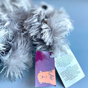 May include: A purple and orange tag with a bear illustration and the text "Chill Fred" attached to a gray plush toy. The tag includes safety information in French and German.