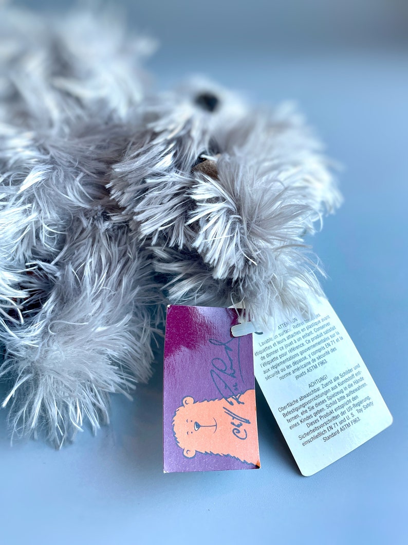 May include: A purple and orange tag with a bear illustration and the text "Chill Fred" attached to a gray plush toy. The tag includes safety information in French and German.