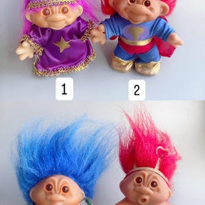 May include: Four troll dolls with different hair colors and outfits. The first troll has pink hair and is wearing a purple robe with a star and a crescent moon. The second troll has red hair and is wearing a blue superhero costume. The third troll has blue hair and is wearing a plaid shirt and brown pants. The fourth troll has red hair and is wearing a white diaper with a jewel.