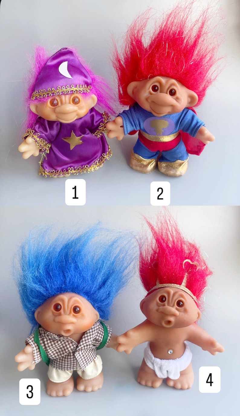 May include: Four troll dolls with different hair colors and outfits. The first troll has pink hair and is wearing a purple robe with a star and a crescent moon. The second troll has red hair and is wearing a blue superhero costume. The third troll has blue hair and is wearing a plaid shirt and brown pants. The fourth troll has red hair and is wearing a white diaper with a jewel.