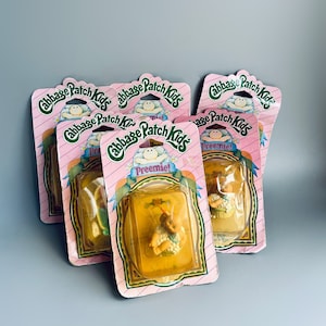 Puede incluir: Seis paquetes sin abrir de muñecas Cabbage Patch Kids Preemie. Cada paquete presenta un fondo rosa y verde con el logotipo de Cabbage Patch Kids y las palabras "Preemie!" en una pancarta amarilla.