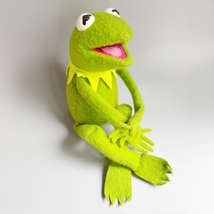 May include: A green felt frog plush toy featuring a bright yellow pointed collar, large white eyes with black pupils, and an open mouth revealing a pink tongue. The figure is depicted in a seated position with long limbs and webbed feet.
