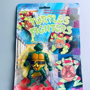 May include: A Teenage Mutant Ninja Turtles action figure in a plastic package. The figure is green with a yellow shell and is holding two katanas. The package has a blue background with the words "TURTLES FIGHTERS" in white letters. The package also has a picture of the Teenage Mutant Ninja Turtles on the front.