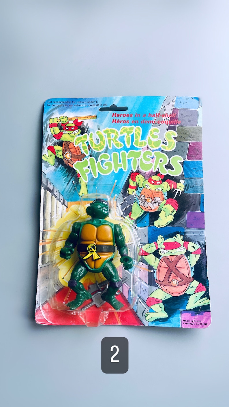 May include: A Teenage Mutant Ninja Turtles action figure in a plastic package. The figure is green with a yellow shell and is holding two katanas. The package has a blue background with the words "TURTLES FIGHTERS" in white letters. The package also has a picture of the Teenage Mutant Ninja Turtles on the front.