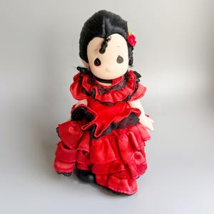 Könnte beinhalten: Puppe in einem roten Rüschenkleid mit schwarzer Spitzenborte, einem schwarzen Halsband und einer roten Blume in ihrem schwarzen Haar. Die Puppe hat einen ruhigen Ausdruck und ist vor einem grauen Hintergrund platziert.