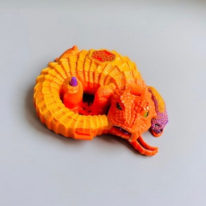 Op de afbeelding: Oranje en paars plastic speelgoed met een krokodilkop en een gekrulde staart. Het speelgoed heeft een rood en gouden logo met de tekst "MM" op de achterkant.