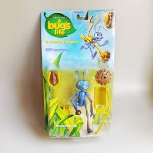 May include: A Disney Pixar A Bug's Life Inventor Flik action figure in original packaging. The blue ant character is accompanied by yellow and brown plastic accessories, including a catapult. The card features the movie logo and text.
