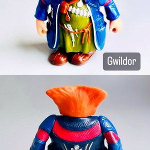 May include: A plastic action figure of a fantasy creature with orange hair, a blue robe, and a green tunic. The figure is standing with its arms at its sides. The figure is labeled "Gwildor".