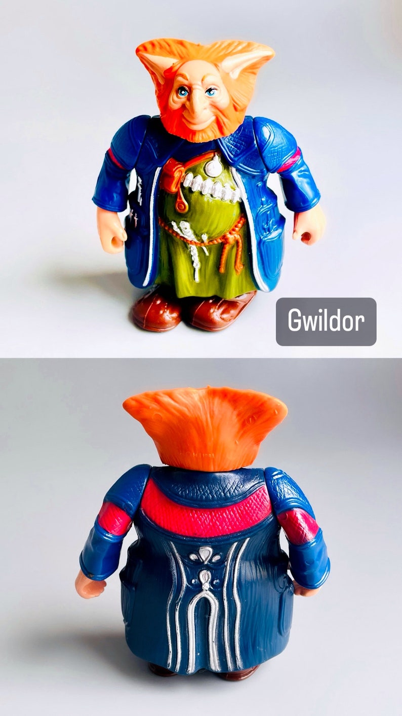 May include: A plastic action figure of a fantasy creature with orange hair, a blue robe, and a green tunic. The figure is standing with its arms at its sides. The figure is labeled "Gwildor".