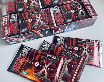 May include: A box of The X-Files trading cards, featuring a red and black design with the show's logo and the text "Season Three". The box is open, revealing packs of cards with the same design. The packs are labeled "Season Three" and "All New".
