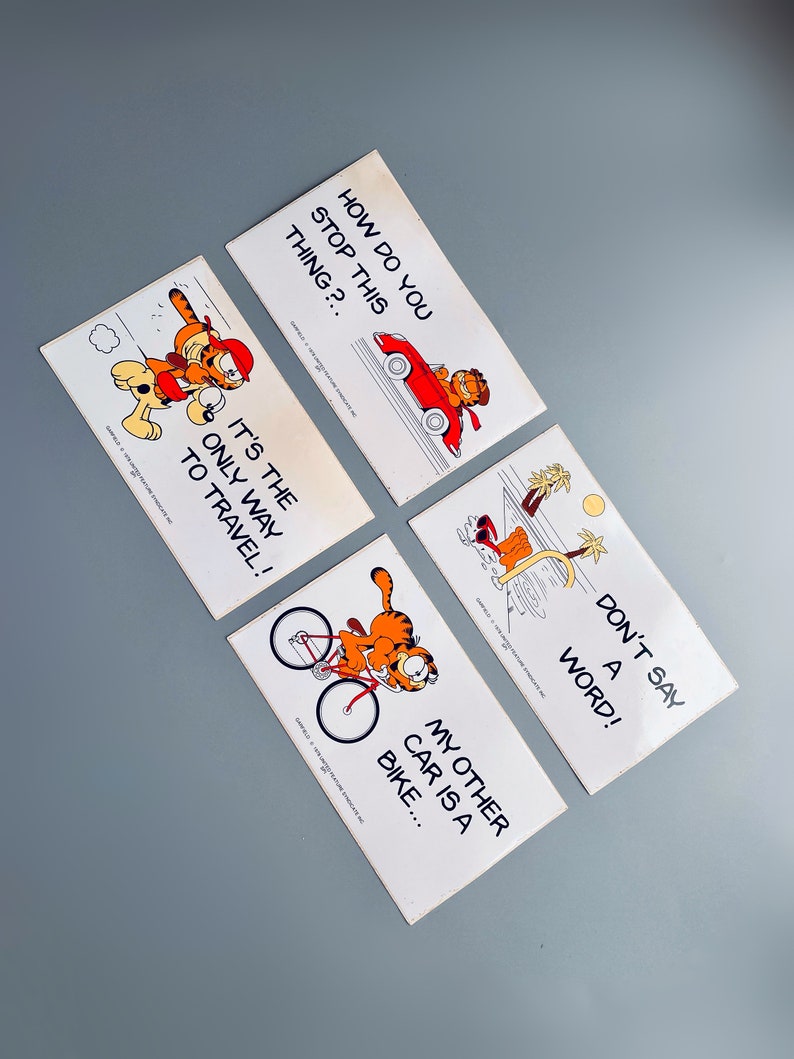 Puede incluir: Cuatro tarjetas blancas con ilustraciones de dibujos animados de Garfield el gato. Las tarjetas presentan diferentes lemas, que incluyen "&iquest;C&oacute;mo se detiene esto?", "&iexcl;Es la &uacute;nica forma de viajar!", "&iexcl;No digas ni una palabra!" y "Mi otro coche es una bicicleta..."