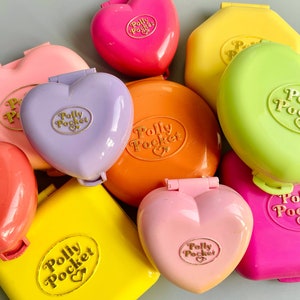 Może przedstawiać: Kolekcja zabytkowych kompaktów Polly Pocket w różnych kolorach, w tym różowym, fioletowym, żółtym, pomarańczowym i zielonym. Kompakty są w kształcie serca i prostokątne, z wytłoczonym logo Polly Pocket z przodu.