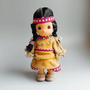 Può includere: Una bambola Precious Moments vestita con abiti da nativo americano. La bambola ha capelli neri intrecciati, una fascia rossa e bianca e un vestito color cachi con rifiniture rosse e blu. La bambola indossa scarpe color cachi e una collana di perline colorate.