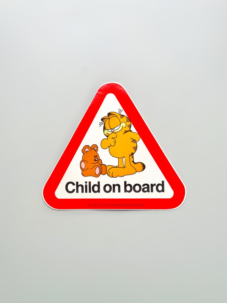 Puede incluir: Pegatina roja en forma de tri&aacute;ngulo con un gato naranja de dibujos animados y un oso de peluche marr&oacute;n. La pegatina dice "Ni&ntilde;o a bordo" con el texto "&copy; 1978 United Feature Syndicate, Inc." en la parte inferior.