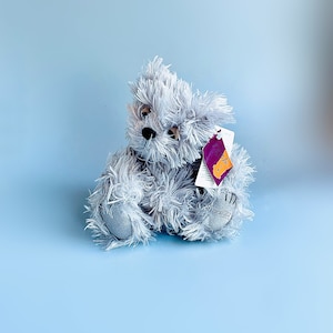May include: A gray plush teddy bear with a purple tag attached to its ear. The bear is sitting on a light blue background.