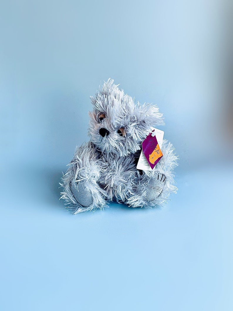 May include: A gray plush teddy bear with a purple tag attached to its ear. The bear is sitting on a light blue background.
