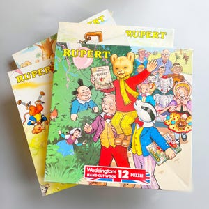 May include: A collection of Rupert the Bear jigsaw puzzles. The boxes display vibrant illustrations of Rupert and his companions. The puzzles are from Waddingtons, with a 12-piece count. The boxes feature the word "RUPERT" in bold yellow lettering.