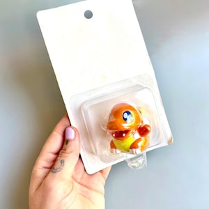 May include: A small orange and yellow plastic toy figure of a Pokemon character in a clear plastic blister pack. The figure is in a standing pose with its arms outstretched.