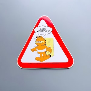Puede incluir: Un cartel de advertencia triangular rojo con un gato naranja de dibujos animados, Garfield, en su interior. El cartel dice "GOOD VIBRATIONS" en una burbuja de pensamiento sobre la cabeza de Garfield. El cartel tambi&eacute;n incluye la informaci&oacute;n de copyright: "SP1064 &copy; 1978 United Feature Syndicate Inc. SPI Ltd (0532) 465471".