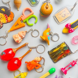 May include: A collection of colourful keychains featuring a variety of themes, including a cartoon bear, a pair of scissors, a bird, a maraca, a monkey, a hamburger, a dog, a pair of shoes, and a small hourglass.