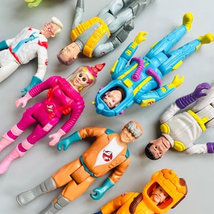 Op de afbeelding: Een verzameling vintage actiefiguren met personages in kleurrijke uniformen. De figuren hebben gedetailleerde gezichten en liggen. Het kleurenpalet omvat oranje, blauw, roze en geel, met het Ghostbusters-logo op sommige.