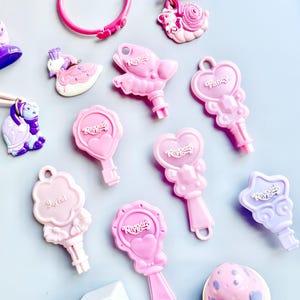 以下が含まれることがあります： ハート、星、白鳥など、さまざまなデザインのピンクとパープルのプラスチック製おもちゃの鍵のコレクション。鍵には「Keypers」という文字が印刷されています。他にも、リング、カタツムリ、ブロックなど、小さなプラスチック製のおもちゃがあります。