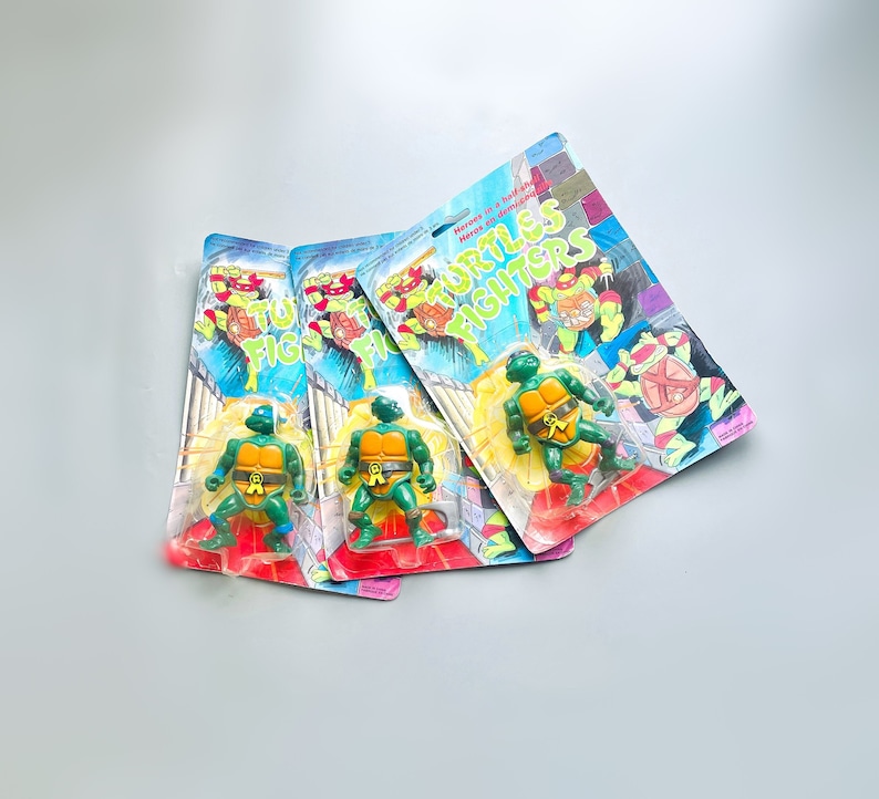 May include: Three Teenage Mutant Ninja Turtles action figures in their iconic green and orange color scheme. The figures are packaged on cardboard cards with the words "Heroes in a Half Shell" and "Teenage Mutant Ninja Turtles Fighters" printed on them.