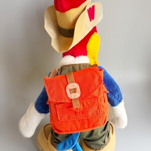 May include: A plush toy of a cartoon bird character, wearing a red hat, tan cowboy hat, and an orange backpack. The bird has blue arms and legs, white gloves, and tan shoes. The hat has a red "W" on the side.
