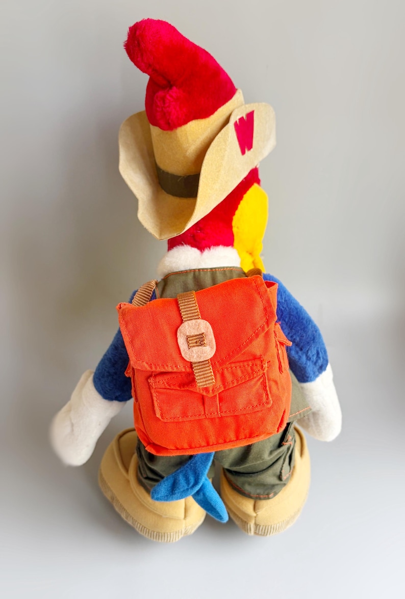 May include: A plush toy of a cartoon bird character, wearing a red hat, tan cowboy hat, and an orange backpack. The bird has blue arms and legs, white gloves, and tan shoes. The hat has a red "W" on the side.