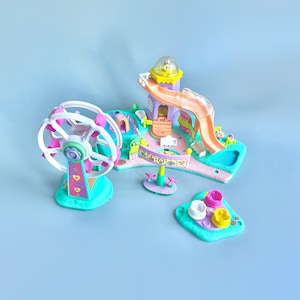 Puede incluir: Un colorido juego de plástico con una noria, un tobogán y un carrusel. El juego está diseñado para muñecas o figuritas pequeñas y presenta un esquema de colores rosa, azul y amarillo brillante.