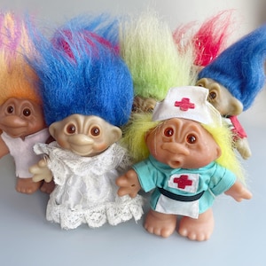 May include: A group of five troll dolls with colorful hair. The dolls are wearing different outfits, including a white dress, a blue dress with a red cross, and a pink dress.