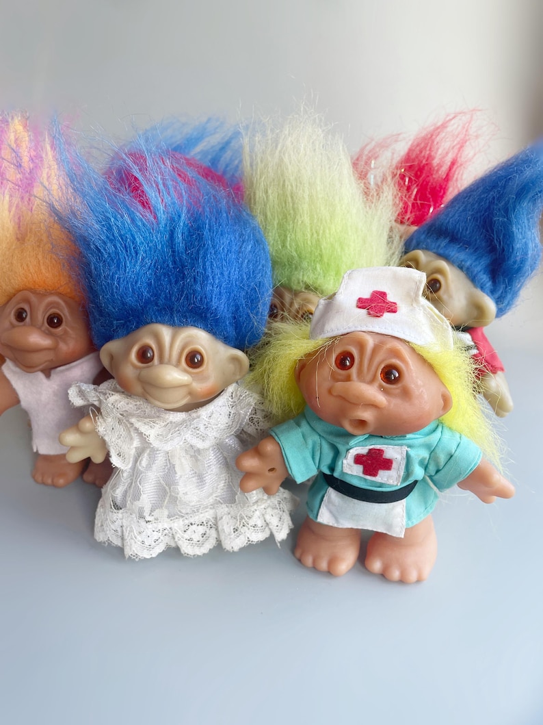May include: A group of five troll dolls with colorful hair. The dolls are wearing different outfits, including a white dress, a blue dress with a red cross, and a pink dress.