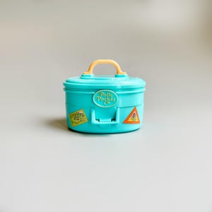 May include: A teal Polly Pocket carrying case with a yellow handle and accents. The case has a rounded shape with a hinged front and is decorated with stickers. The Polly Pocket logo is visible on the front.