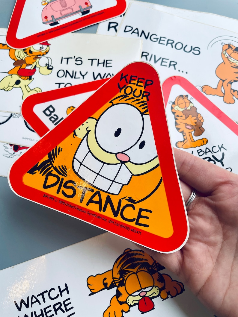 Puede incluir: Un cartel de advertencia triangular rojo y amarillo con una cara de gato de dibujos animados y el texto "KEEP YOUR DISTANCE". El cartel es una pegatina de Garfield con la informaci&oacute;n de copyright "SPI 070 &copy; 1978 United Feature Syndicate Inc. SPI Ltd (0532) 465471."
