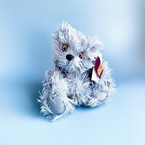 May include: A gray plush teddy bear with a tag attached to its ear. The bear is sitting on a light blue background.